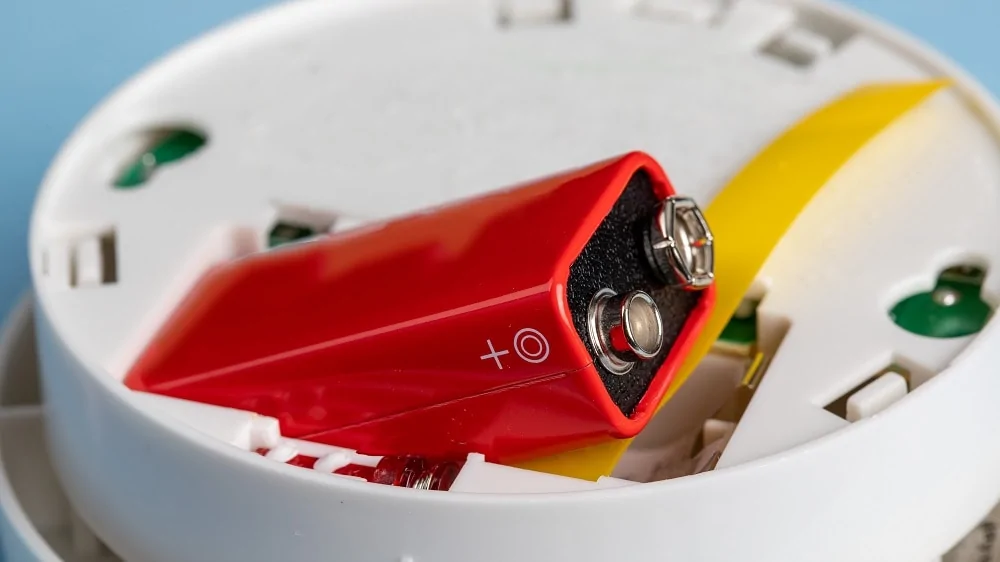 de achterkant van een witte koolmonoxidemelder waaruit een rode alkine batterij steekt