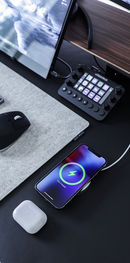 IPhone die draadloos oplaadt op een bureau met ernaast een muis en  een monitor en eronder een case van draadloze oordoppen