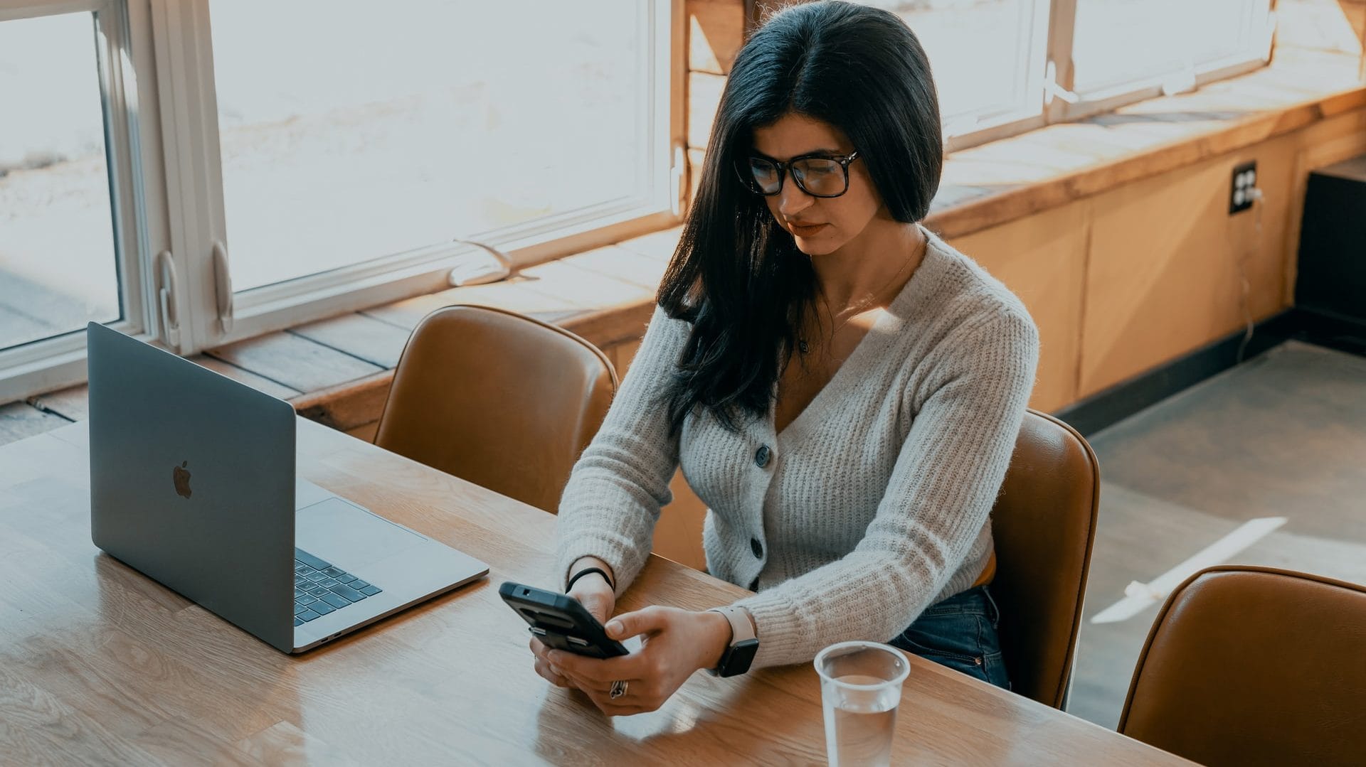 Vrouw in grijze trui zit achter laptop aan tafel met mobiel in hand