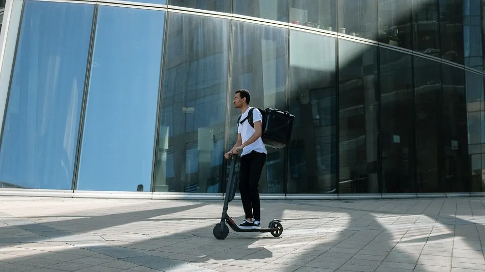 Man in wit shirt met een bezorgtas op zijn rug rijdt op een zwarte elektrsiche scooter voor een glazen gebouw