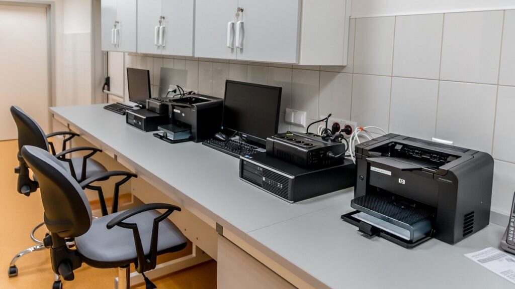 Lang wit bureau waarop meerdere zwarte printers, computermonitoren met toetsenborden staan. Twee bureaustoelen staan aan het bureau en boven het bureau hangen een aantal witte kastjes