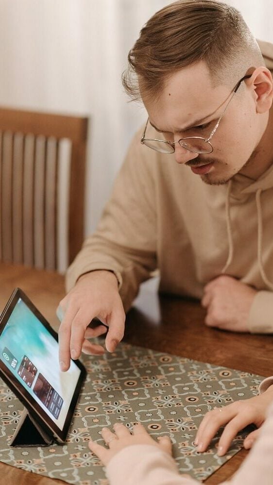 Man met bril in beige trui kijkt verward naar het scherm van een zwarte iPad