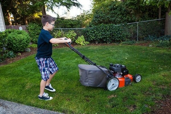 Kleine jongen maait het gras met een rode elektrische grasmaaier met opvangzak
